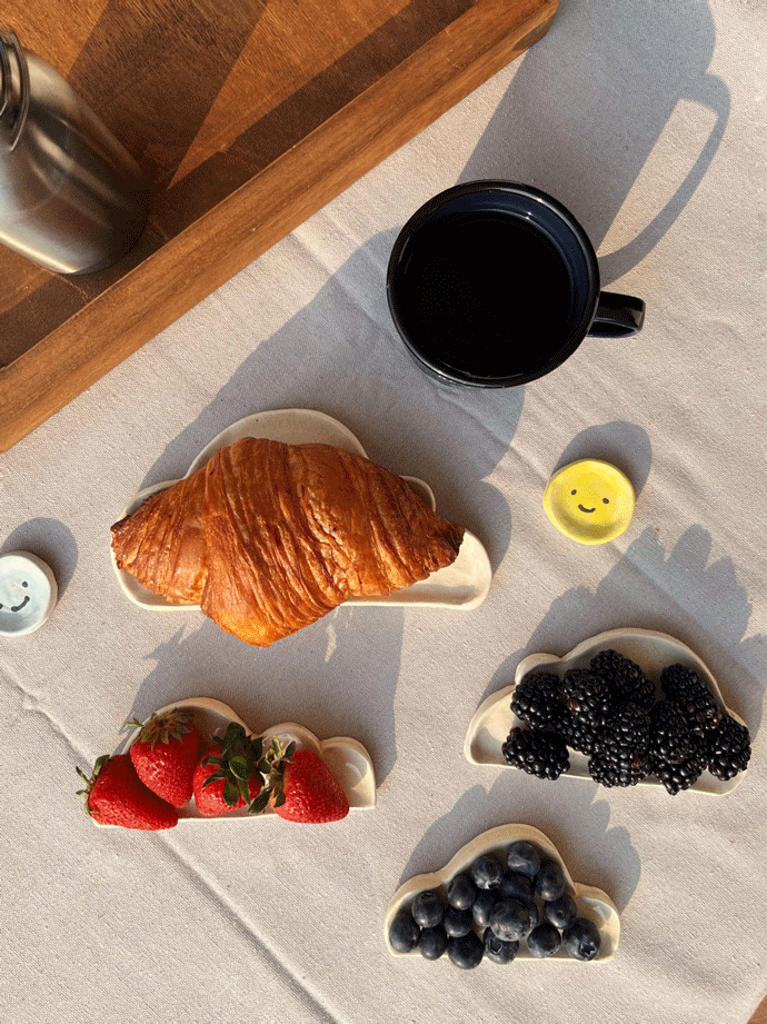 Platos de nubes de cerámica con fruta y croissants encima de una mesa con mantel.